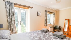 A bedroom with a bed, a chair, a mirror, and a window with curtains. - The Meadows Cottage (Dereham)