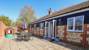 A brick house with a tiled roof, a patio with a table and chairs, and a small playhouse. - The Meadows Cottage (Dereham)
