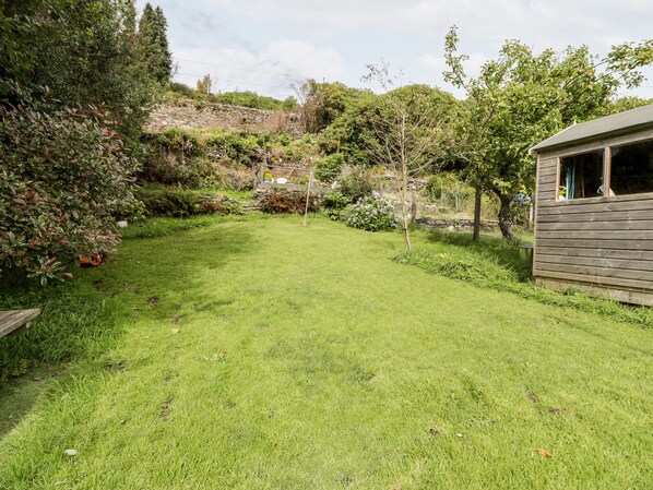 A grassy backyard with a wooden shed, stone wall, and various plants. - Ty Gwynn (Blaenau Ffestiniog)