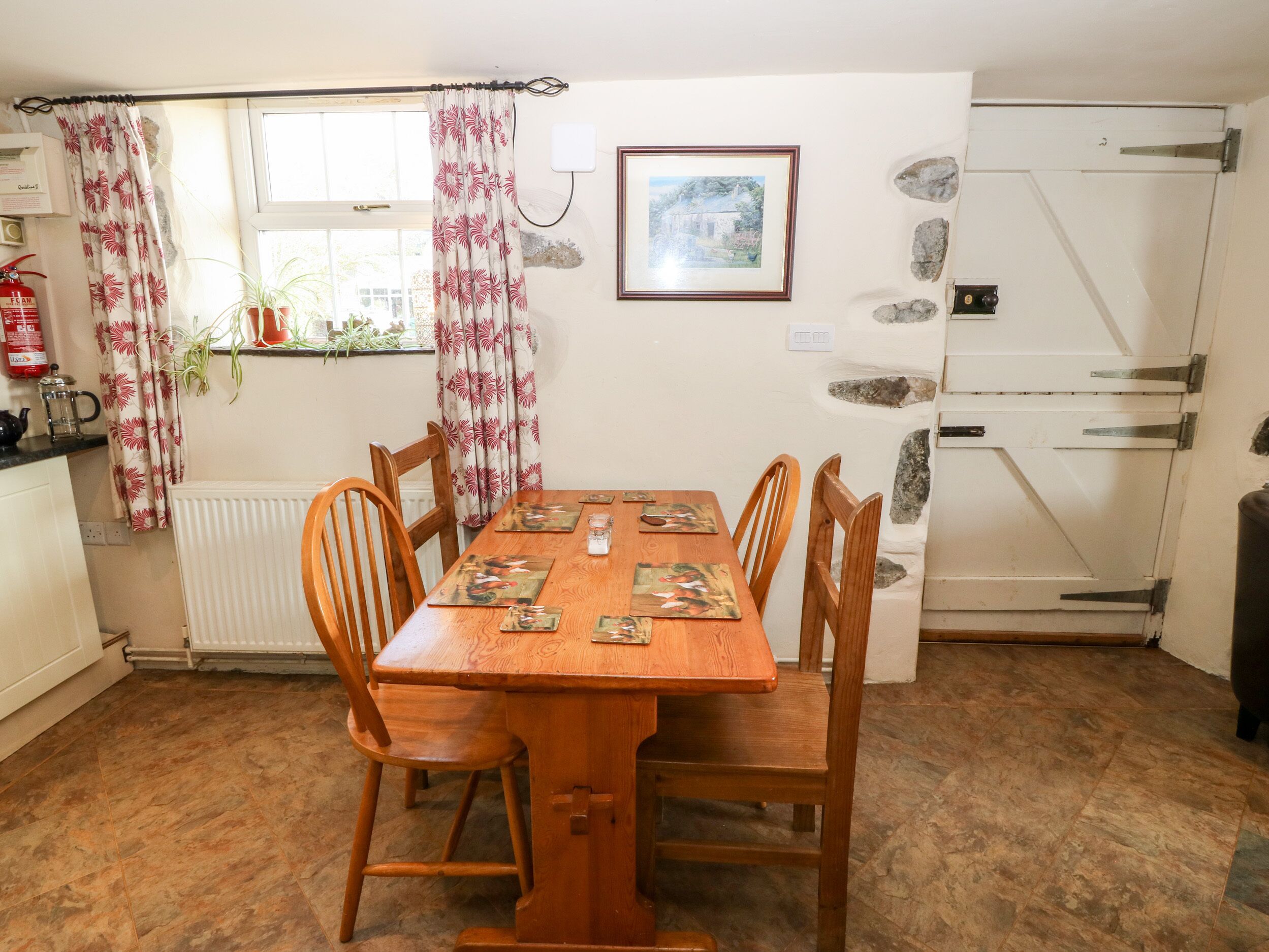 A dining area with a wooden table and chairs, a window with curtains, a radiator, a fire extinguisher, and a stone wall with a picture.