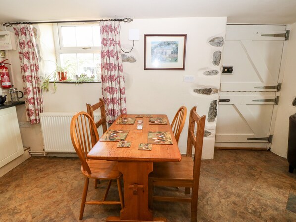 A dining area with a wooden table and chairs, a window with curtains, a radiator, a fire extinguisher, and a stone wall with a picture. - Bwthyn Celyn (Pwllheli)