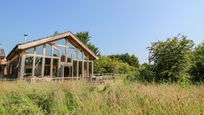 A wooden cabin with large windows and a sloped roof, surrounded by tall grass and trees. - The Tractor Shed (Shrewsbury)