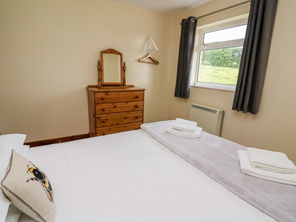 A bedroom with a bed, a wooden dresser, a mirror, and a window with curtains. - Red Kite Cottage (Llandrindod Wells)
