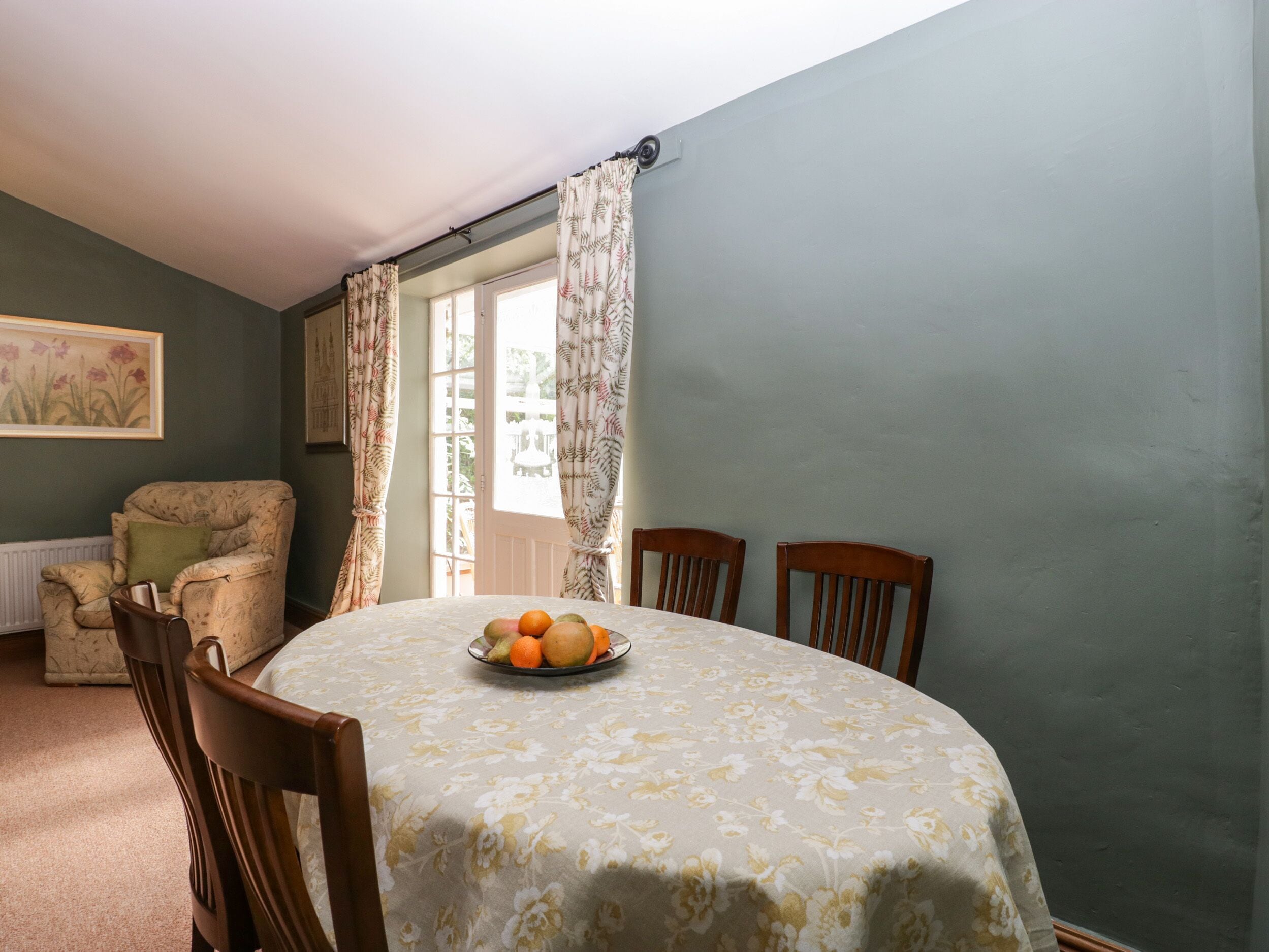 Un coin repas avec une table ronde recouverte d’une nappe à motifs floraux, deux chaises en bois, un fauteuil à motifs floraux et une fenêtre avec des rideaux.