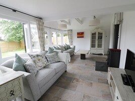 A spacious living room with a grey sofa set, a coffee table, and a TV stand. There is a fireplace and a large window with curtains.