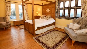 Una cama con dosel con cabecera de madera, una mesita de noche con lámpara, una silla con estampado floral y un sofá con manta de piel.