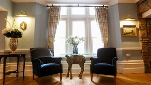 Una habitación con dos sillones azul oscuro, una mesita con un florero y una estatua decorativa de un ciervo, además de un amplio ventanal con cortinas.