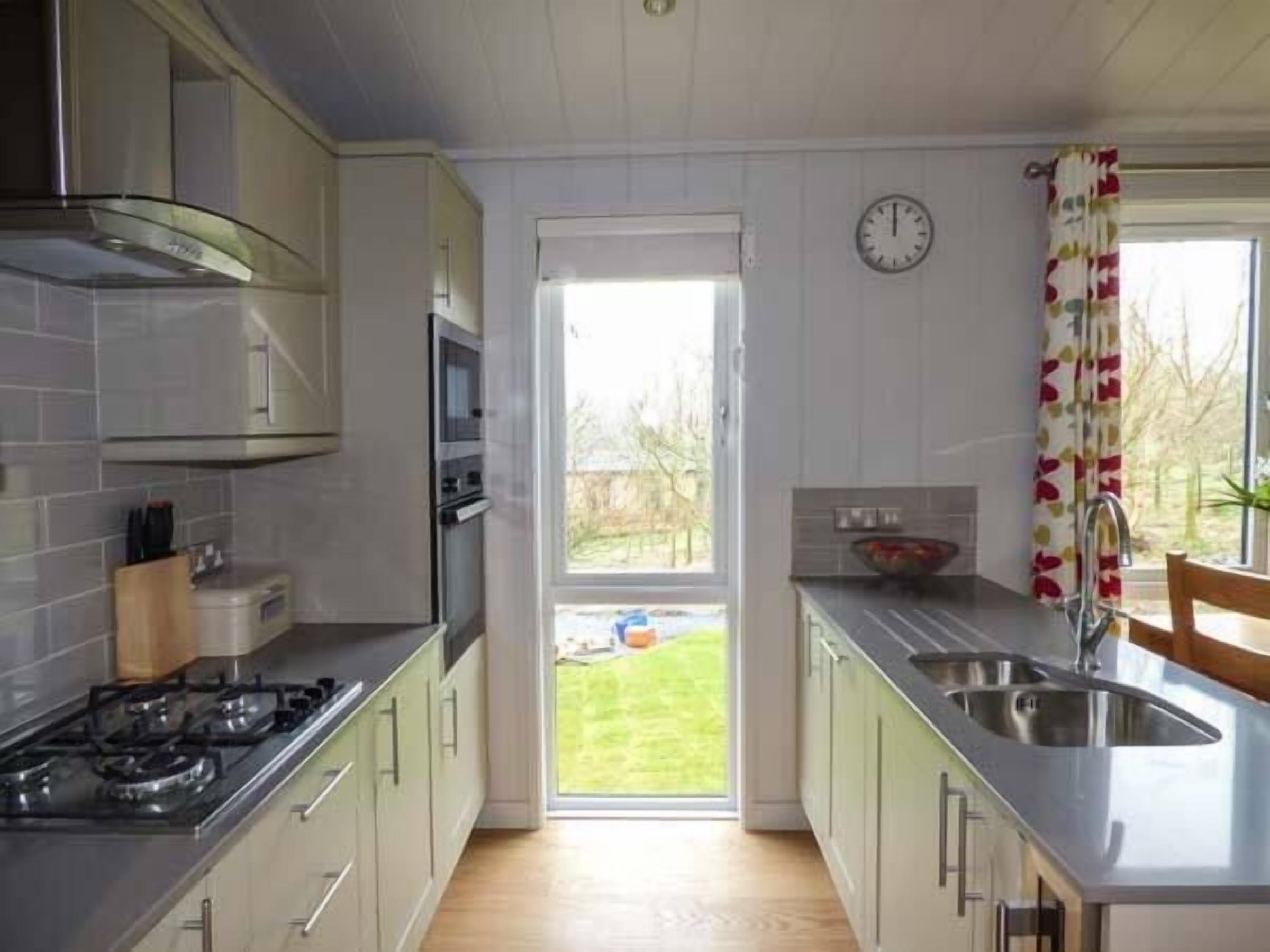 Una cucina moderna con piano cottura a gas, forno e lavello in acciaio inossidabile. Si vede un giardino dalla finestra, e c'è un orologio appeso al muro.