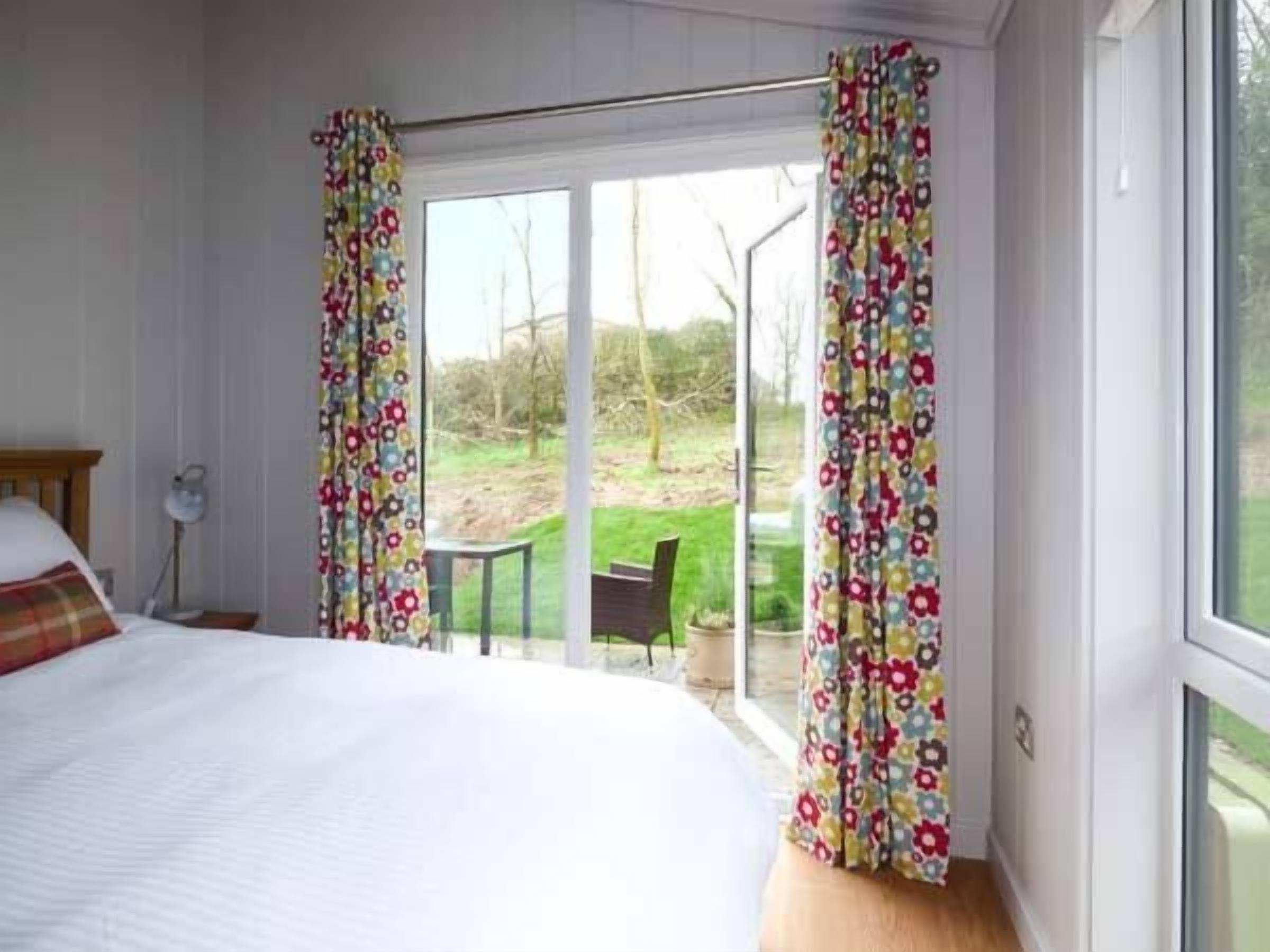 A bedroom with a large bed, a desk, and a chair. There is a sliding glass door leading to a garden.