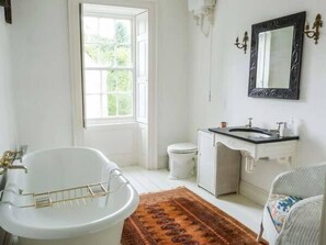 Une salle de bain avec une baignoire sur pieds, des toilettes, un lavabo doté d’un plan de travail noir et un miroir au-dessus du lavabo. Une fenêtre laisse entrer la lumière naturelle et un tapis à motifs recouvre le sol.