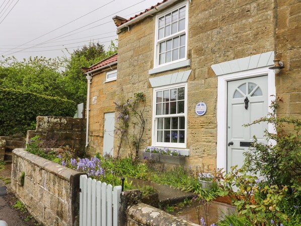 A stone cottage with a white door, a sign reading "Belle Vue Cottages," and a garden with purple flowers. - 1 Belle Vue (Northallerton)