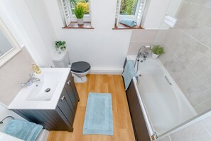 A compact bathroom with a sink, toilet, and bathtub. There are two windows with plants, a blue towel on the floor, and a small shelf with a plant. - 1 Belle Vue (Northallerton)