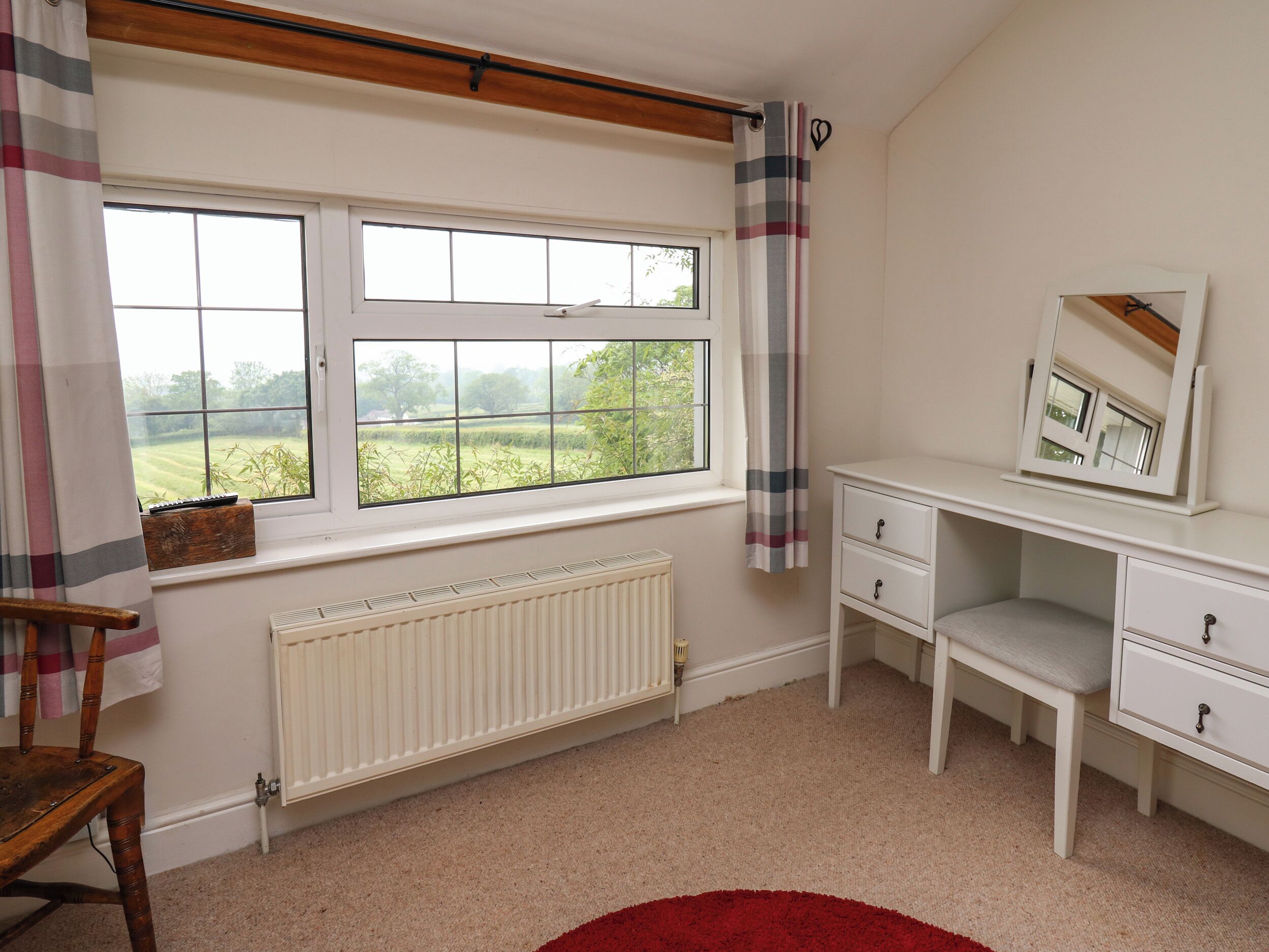 A room with a large window, a radiator, a desk with drawers, a chair, and a red rug.