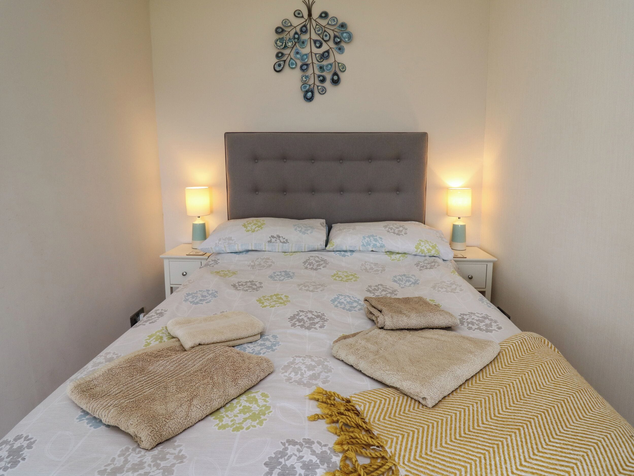 Uma cama bem arrumada com edredom estampado, duas luminárias de cabeceira e um objeto decorativo na parede acima da cama.