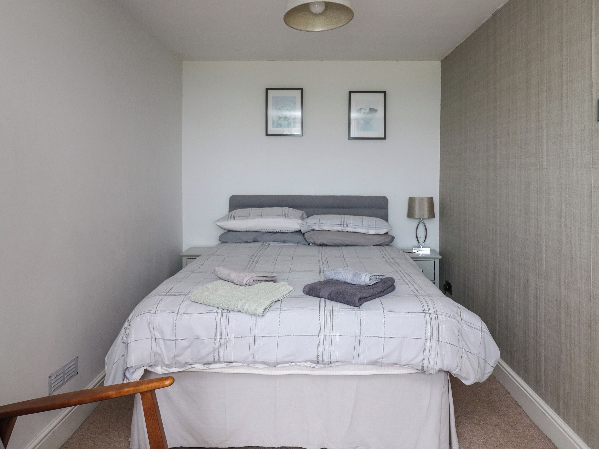 Una cama bien hecha con una colcha de cuadros grises y blancos, dos almohadas y toallas dobladas sobre la cama. Hay una mesita de noche con una lámpara y un cuadro enmarcado en la pared sobre la cama.
