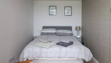 Una cama bien hecha con una colcha de cuadros grises y blancos, dos almohadas y toallas dobladas sobre la cama. Hay una mesita de noche con una lámpara y un cuadro enmarcado en la pared sobre la cama.