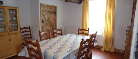 A dining room with a long table, wooden chairs, a wooden cabinet with glass doors, a wooden door, and a window with yellow curtains.