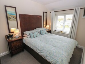 Une chambre à coucher bien rangée, avec un grand lit, des lampes de chevet, une table de nuit, une commode, une fenêtre avec des rideaux et un tableau encadré accroché au mur.