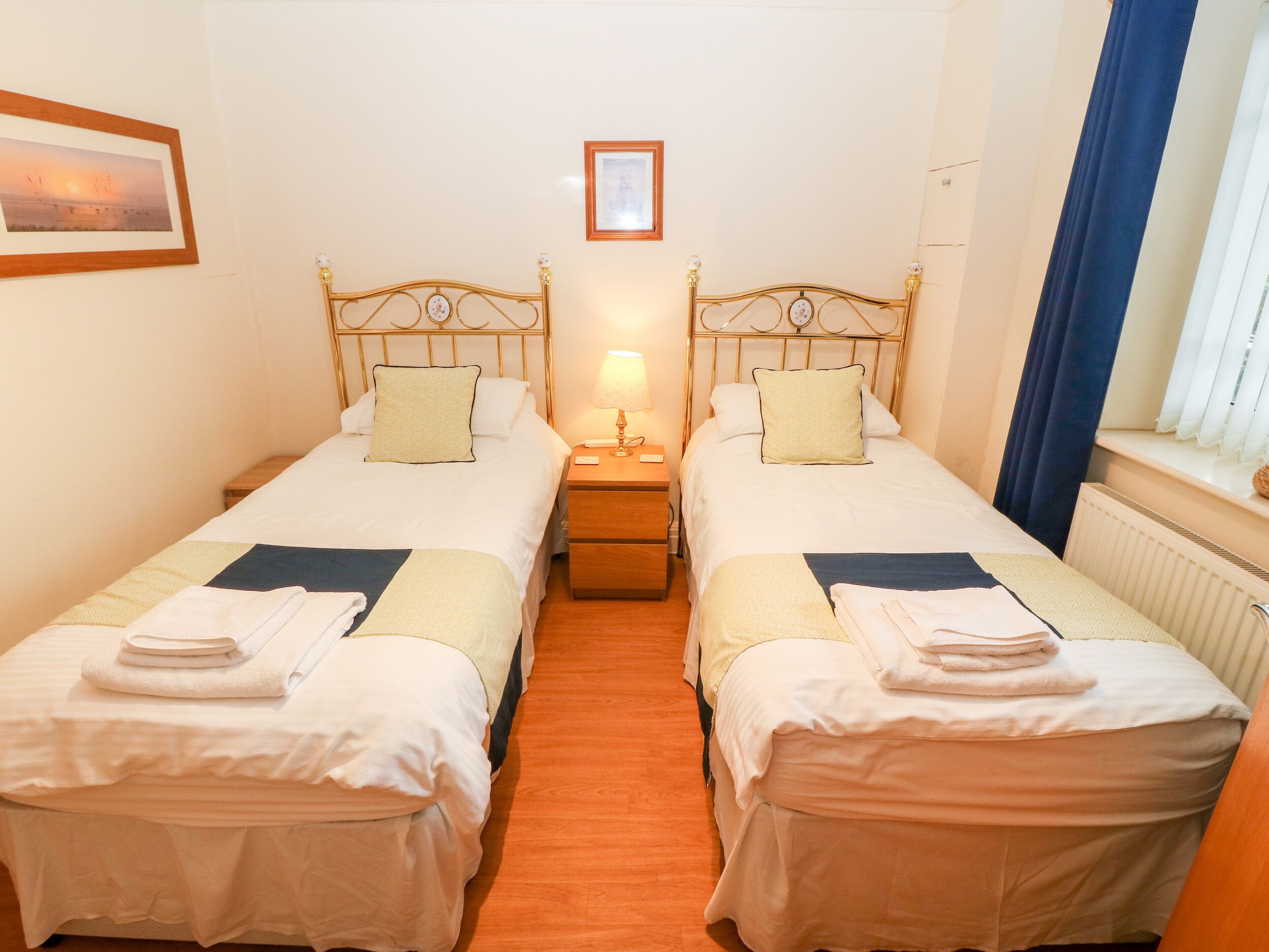 A twin room with two single beds, a nightstand with a lamp, a framed picture on the wall, and a window with curtains.