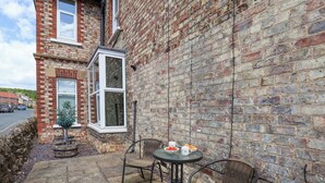 Ein Backsteinhaus mit kleiner Terrasse, ausgestattet mit Tisch, Stühlen, einem Blumentopf und einem gepflasterten Weg.