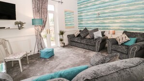 A living room with a grey sofa, teal cushions, a floor lamp, and a painting of a landscape. - St Cuthbert's Cottage (Berwick-upon-Tweed)