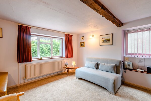 A cozy room with a sofa, a chair, a radiator, a window with curtains, and a small table with a lamp. - The Olde House (Ross-on-Wye)