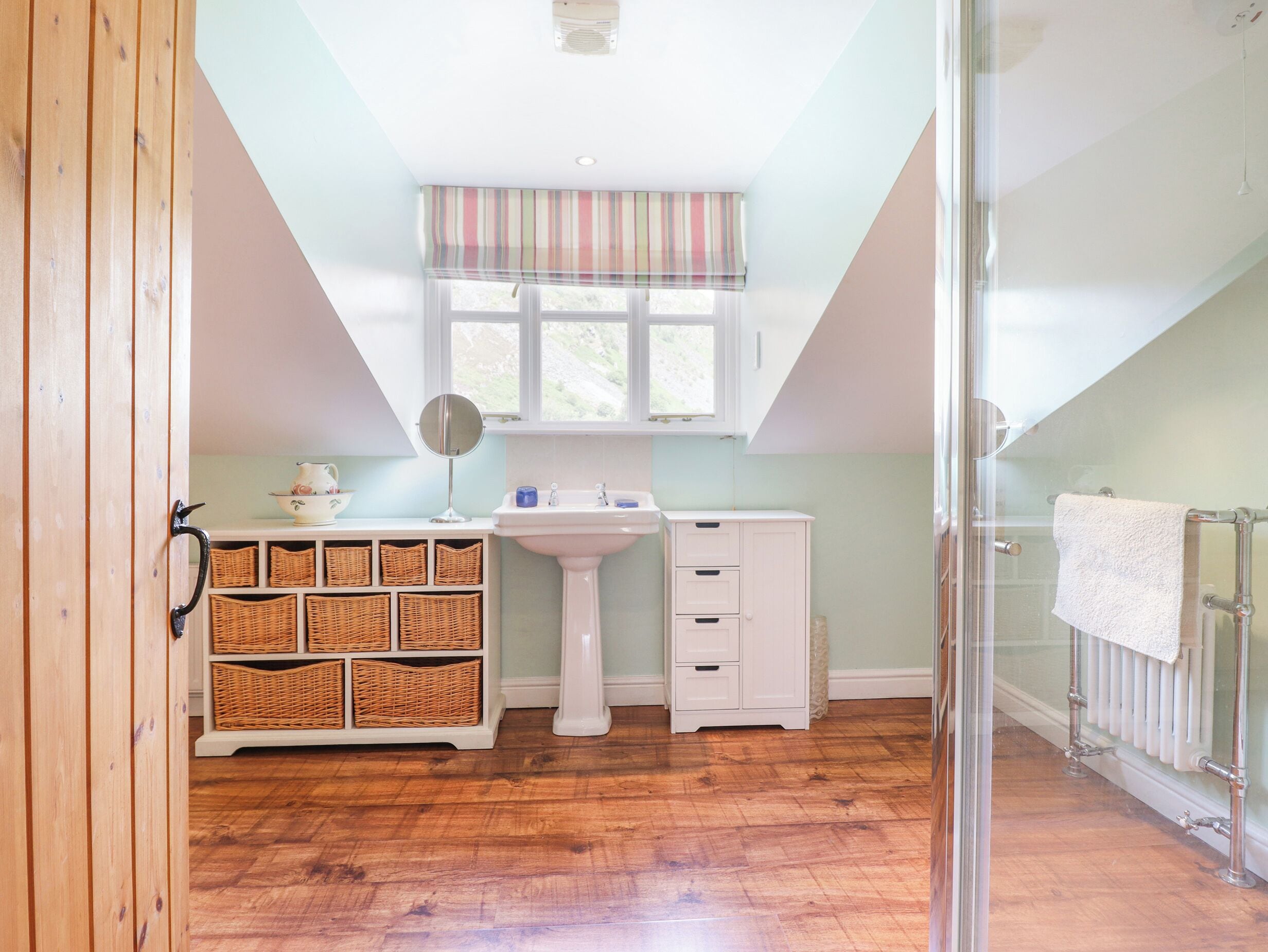 A small, bright room with a wooden floor, a white sink, a mirror, wicker baskets, and a window with a striped valance.
