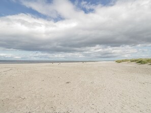 Une vaste plage de sable, bordée de dunes sur la droite, et un ciel nuageux.