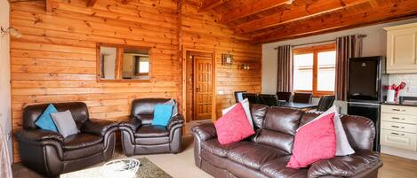 A cozy log cabin interior with a living room featuring leather sofas and armchairs, a dining area with a table and chairs, and a kitchen with a refrigerator and cabinets.