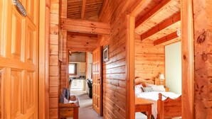 Interior de una cabaña de madera con un estrecho pasillo que conduce a un dormitorio con cama y escritorio.