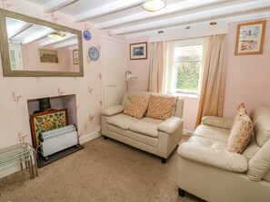 A cozy living room with a beige sofa, a smaller armchair, a radiator, and a window with curtains. - Tir Bedw (Pwllheli)