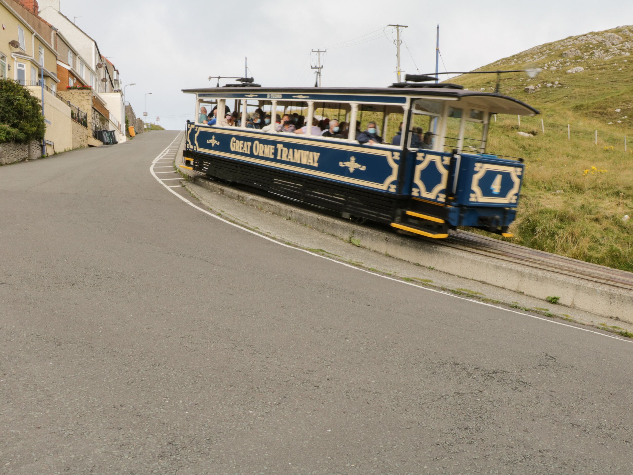 A blue and white tram with "Great Orme Tramway" written on the side, traveling on a road with buildings on one side.