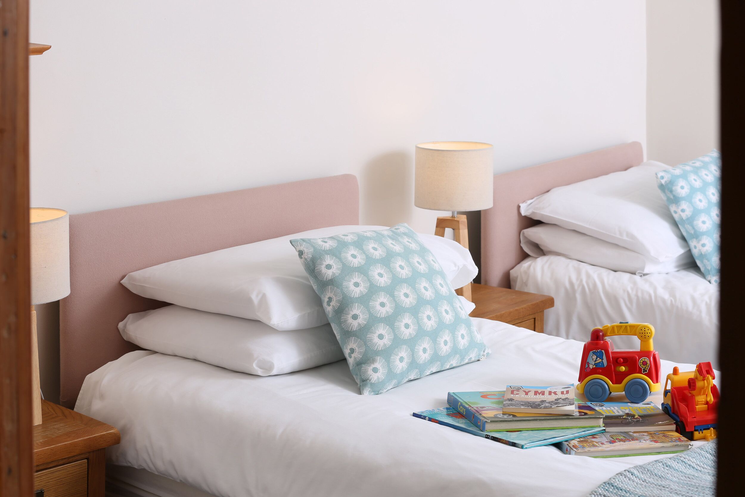 A neatly made bed with white bedding and a blue floral pillow, a small bedside table with a lamp, and a toy fire truck on the bed.