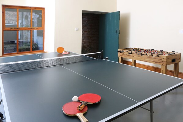 Una mesa de ping pong con dos paletas rojas y una pelota, una mesa de fútbol de mesa y una ventana con vista a un edificio de ladrillo.