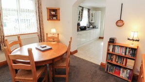 Un coin repas avec une table ronde en bois et des chaises, une bibliothèque remplie de livres, et une vue sur la cuisine à travers une arche.