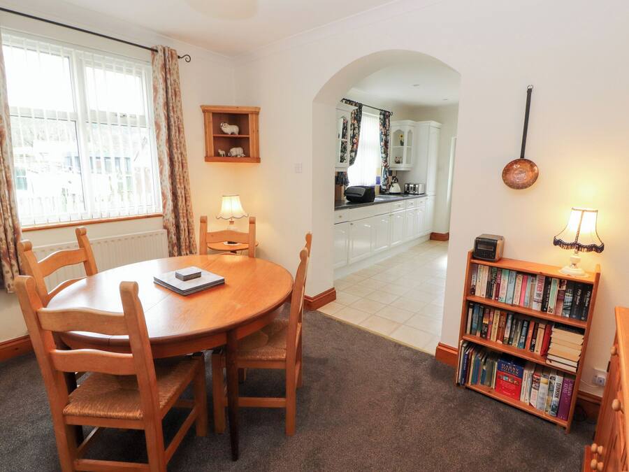 A dining area with a round wooden table and chairs, a bookshelf with books, and a view into the kitchen through an archway.