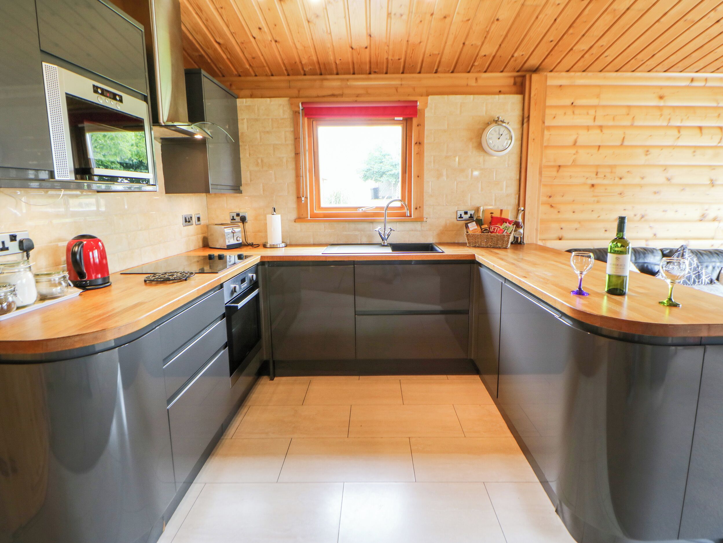 A modern kitchen with a wooden ceiling, a built-in oven, a sink, and a window with a red frame.