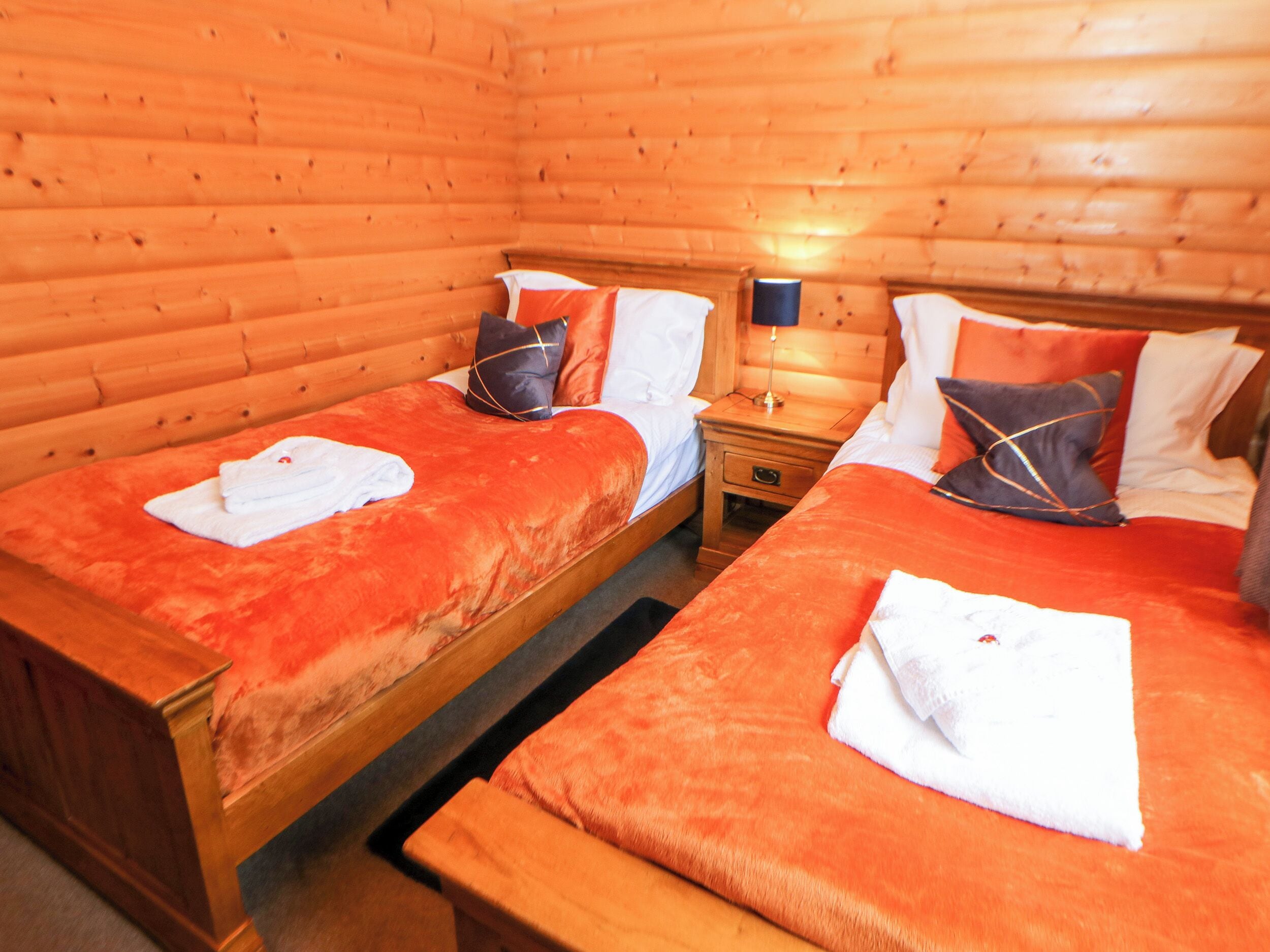 Une chambre avec deux lits en bois, chacun recouvert d’une couverture rouge et agrémenté de serviettes blanches. Une table de chevet avec une lampe se trouve entre les deux lits.