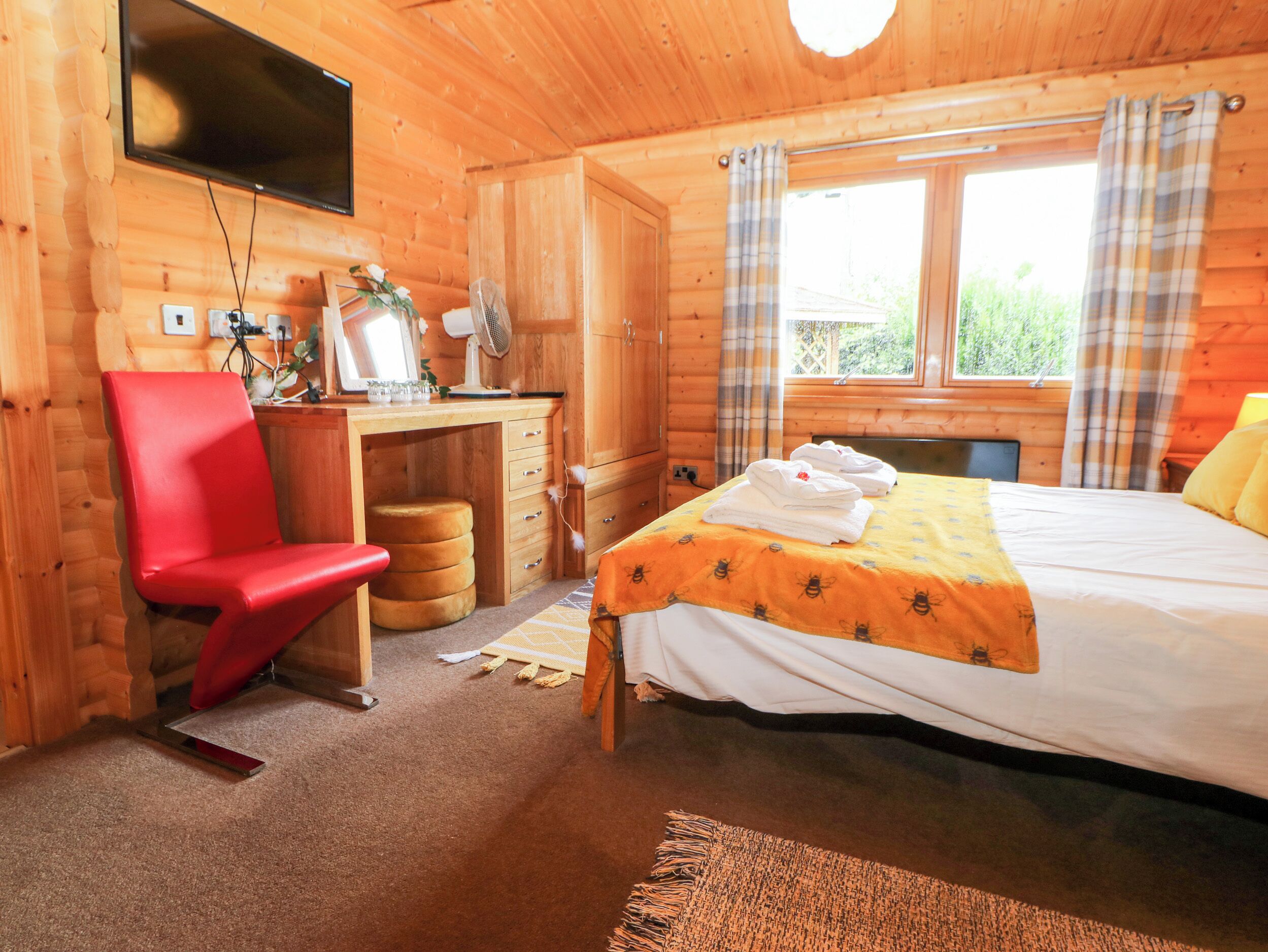 Quarto de cabana de madeira com cama, escrivaninha, cadeira e televisão fixada na parede.