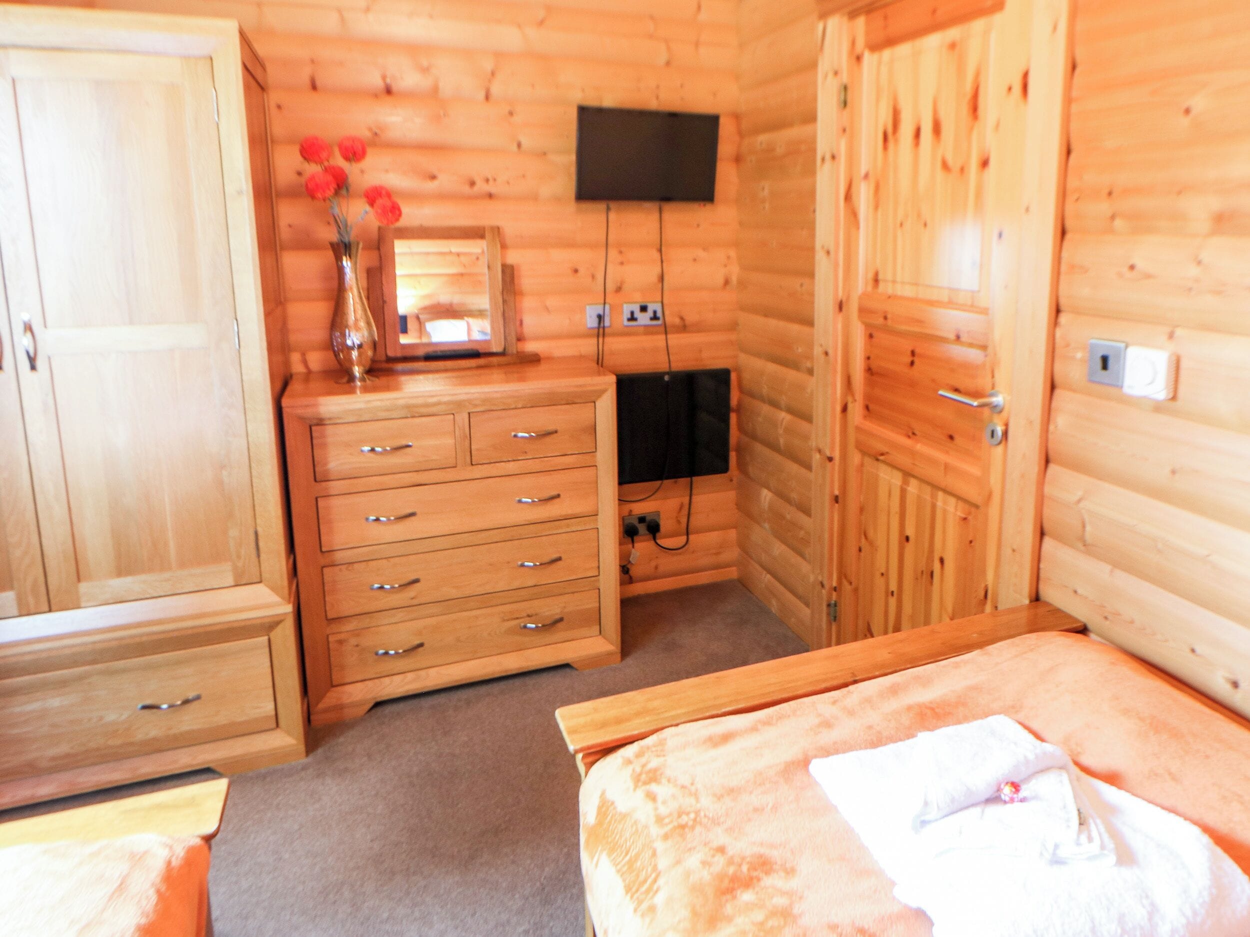 Ein Holzhüttenraum mit einem Bett, einem Schminktisch, einem an der Wand befestigten Fernseher und einem Holzschrank.