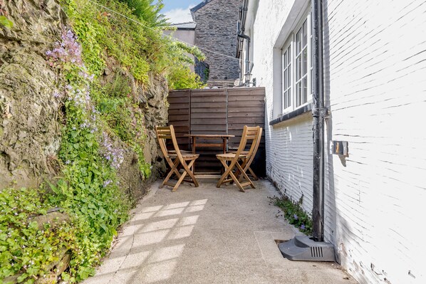A narrow outdoor patio with a wooden table and chairs, surrounded by a white wall and a stone wall with vegetation. - Kynaston Cottage (Aberdovey)