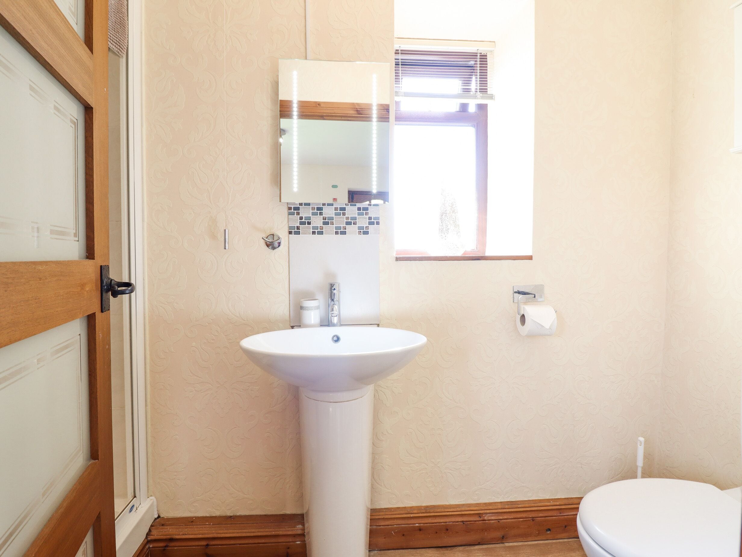 Un baño con un lavamanos blanco, un inodoro, una ventana con persianas y una puerta con marco de madera.