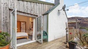 Une terrasse en bois avec une porte-fenêtre coulissante donnant sur une chambre à coucher équipée d’un lit et d’une petite table.