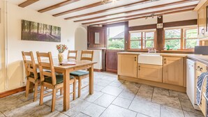 A kitchen with wooden cabinets, a dining table with chairs, and a window with a view of greenery. - West End Barn (Evesham)