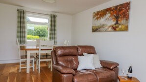A brown leather sofa with a white pillow, a wooden table with two wine glasses and a bottle, a dining table with chairs, and a window with curtains. - Foxes Meadow (Llandrindod Wells)