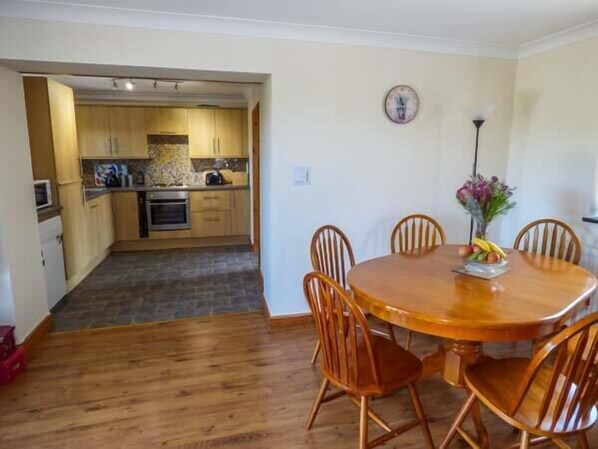 A dining area with a wooden table and chairs, a clock on the wall, and a kitchen in the background. - Ysgubor - Barn (Abergele)