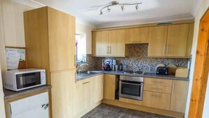 A compact kitchen with wooden cabinets, a microwave, a stove, and a tiled backsplash. - Ysgubor - Barn (Abergele)