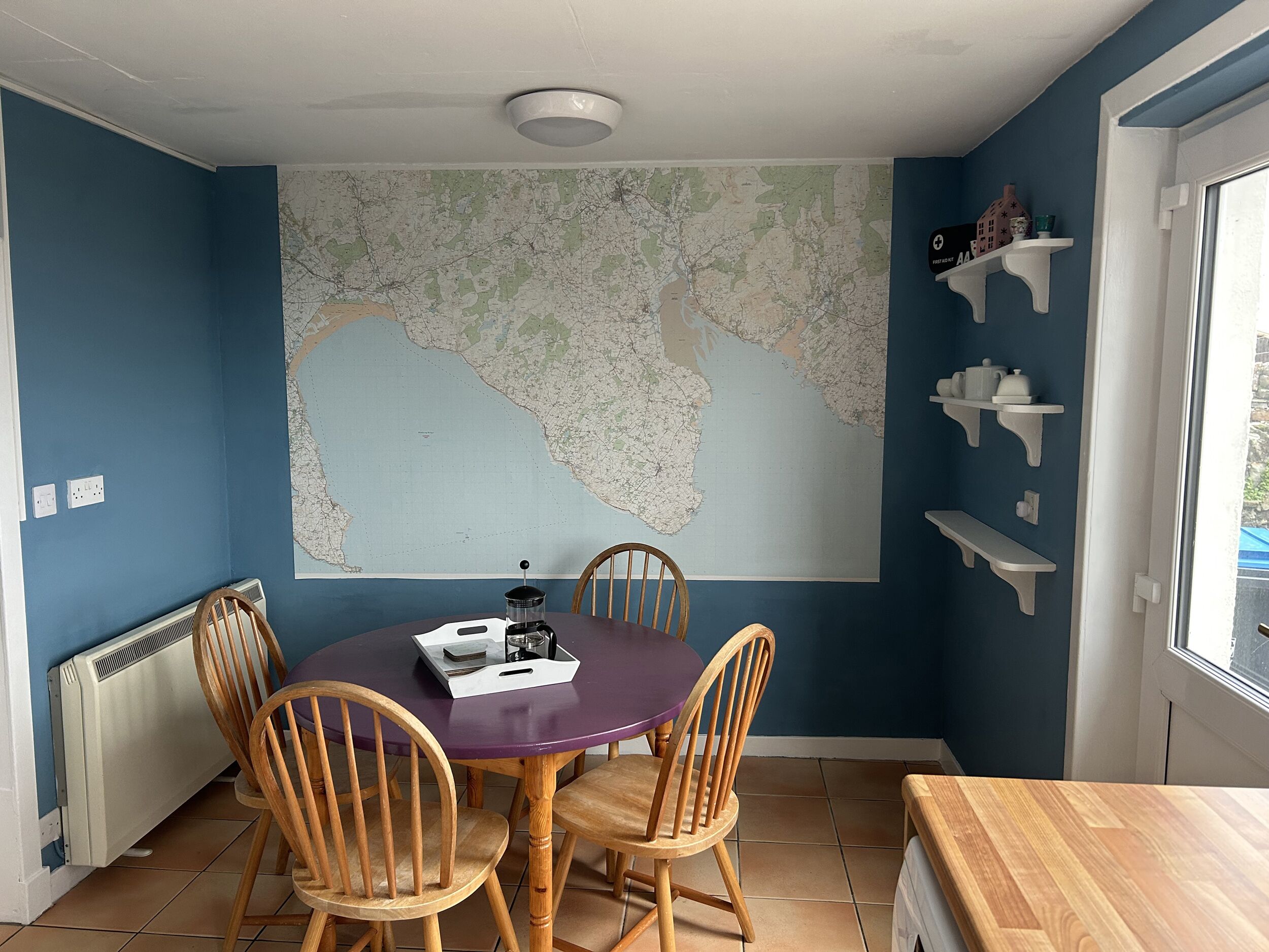 A dining area with a round table, four wooden chairs, a map on the wall, and white shelves with decorative items.