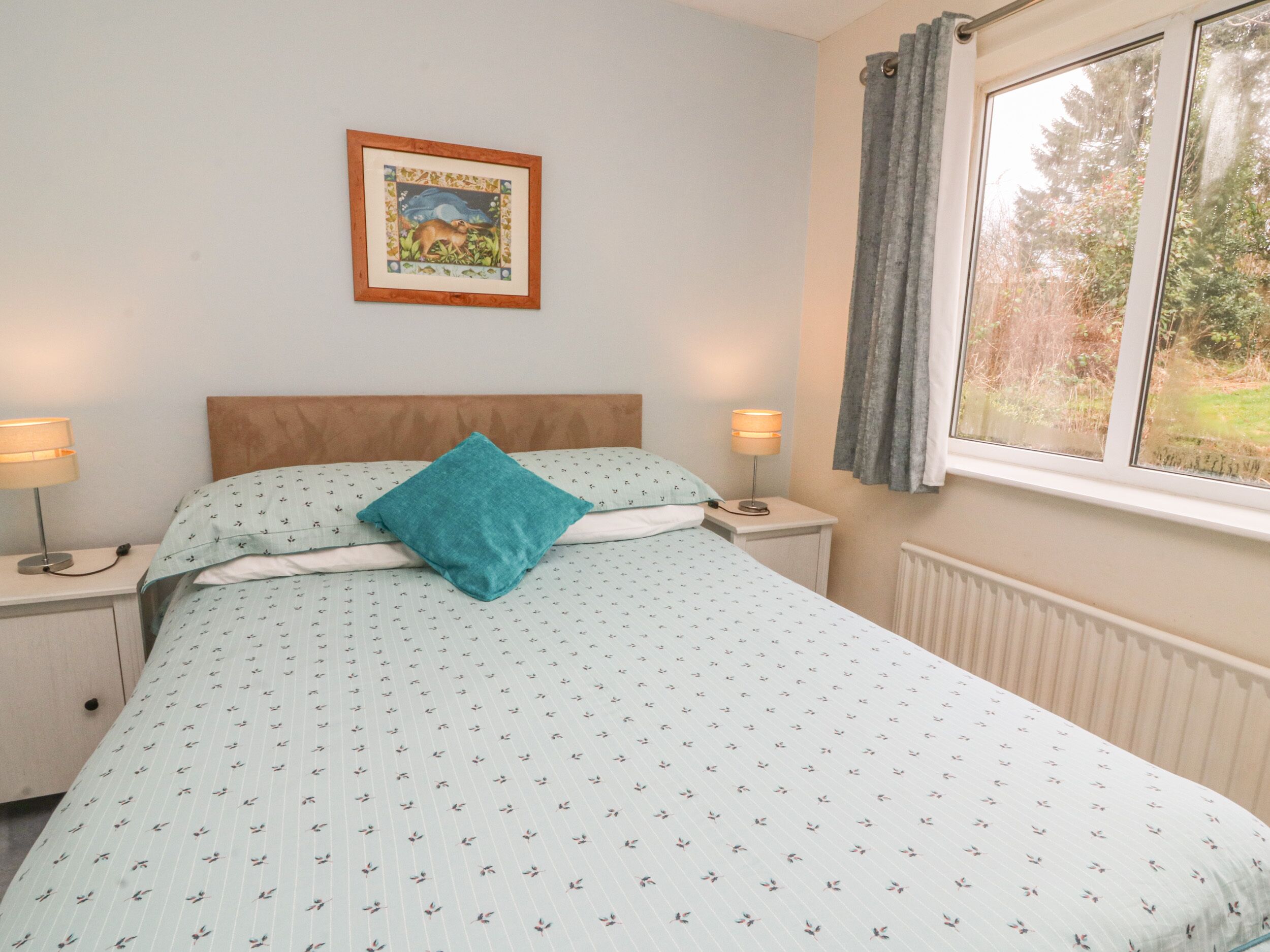 A bedroom with a bed, two bedside tables, a nightstand, a window with curtains, and a framed picture on the wall.