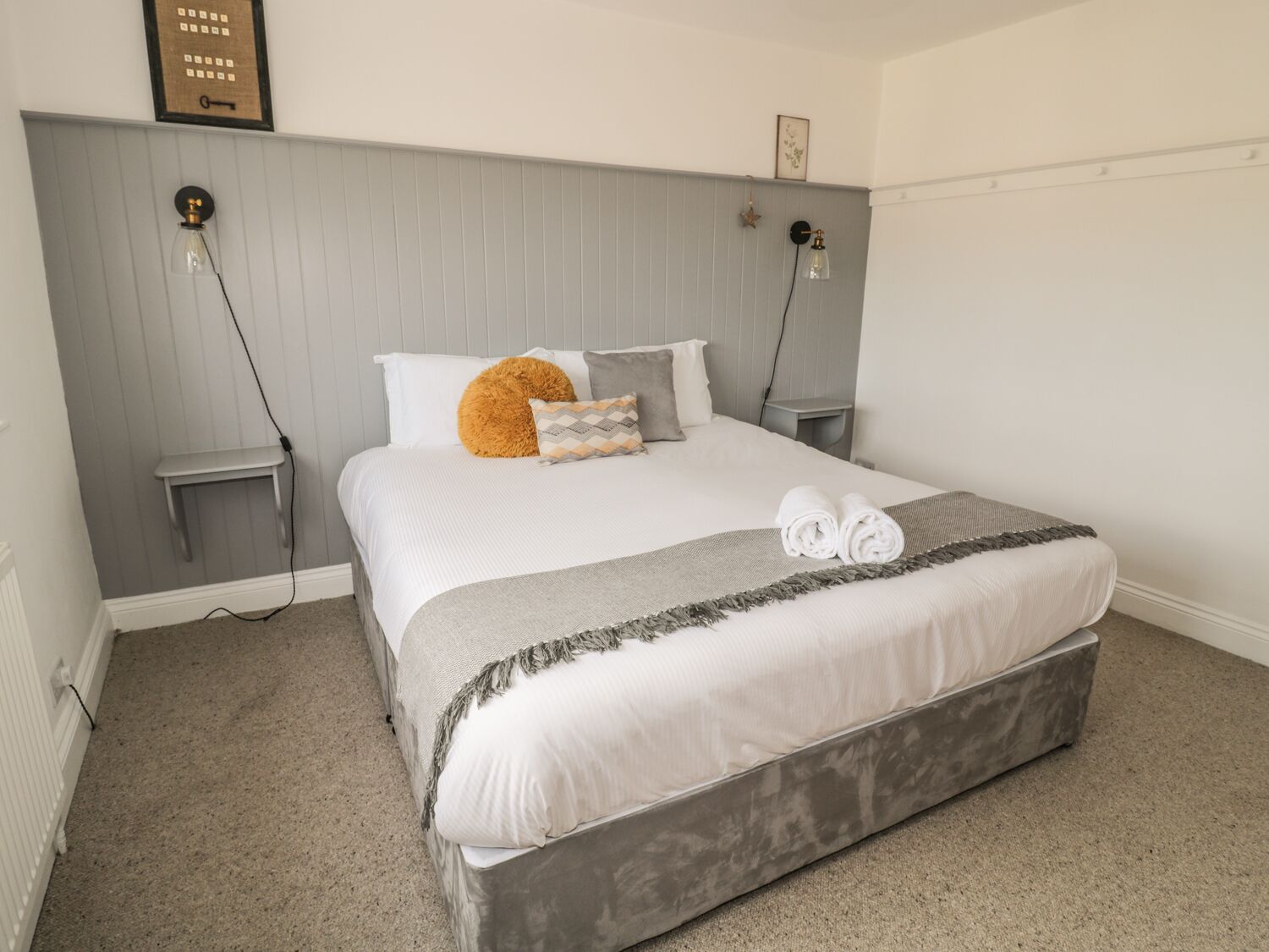 A neatly made bed with a grey headboard, a fluffy orange pillow, and a grey throw blanket. There are two bedside tables with lamps, and a framed picture on the wall.